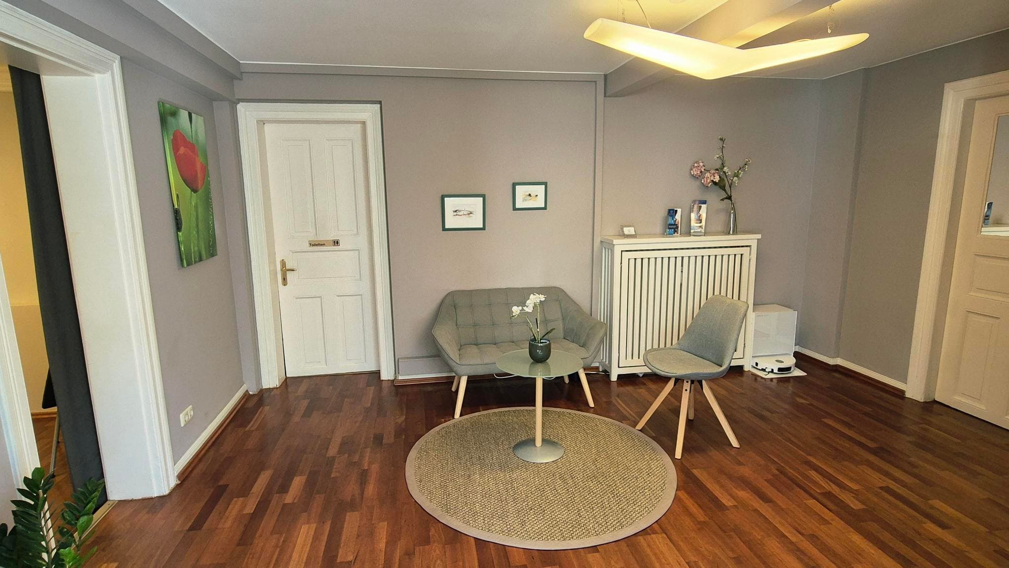 Wartebereich der Praxis mit Holzboden, kleinem Sofa, Stuhl und rundem Tisch mit Orchidee